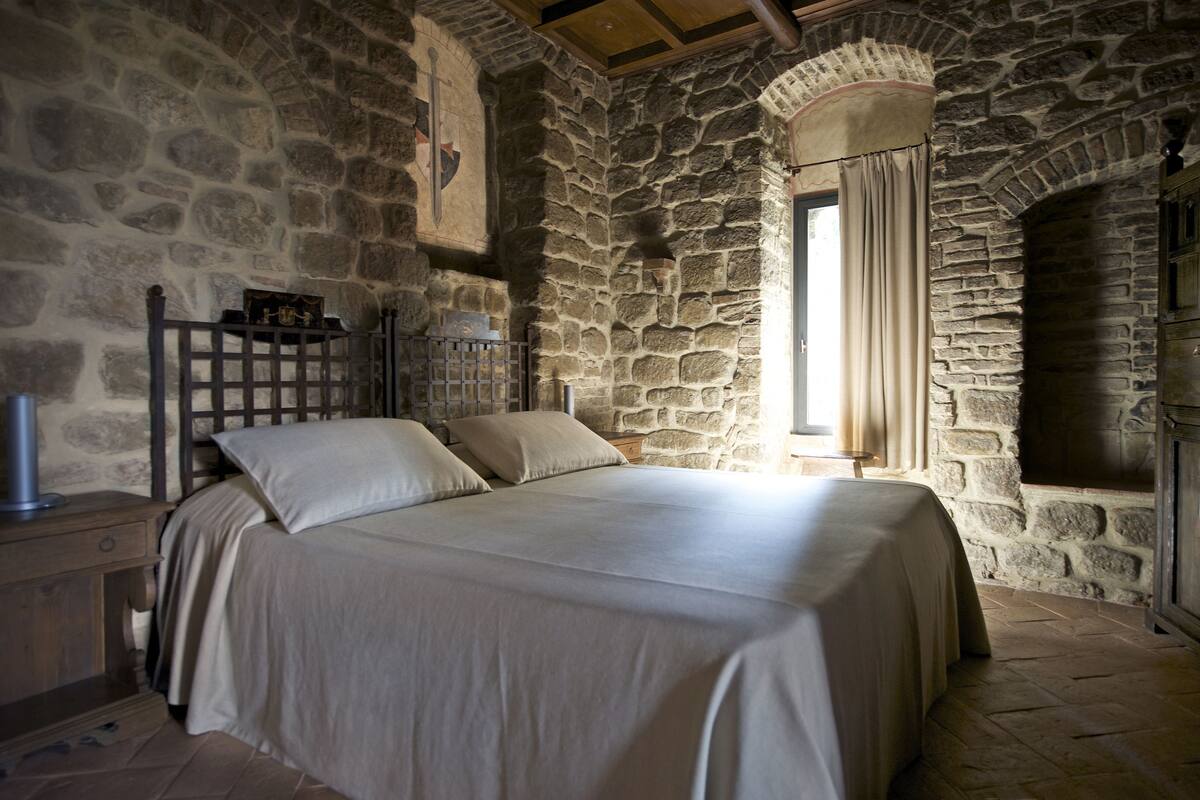 A stone-walled bedroom with a bed, a nightstand, and a window with curtains.