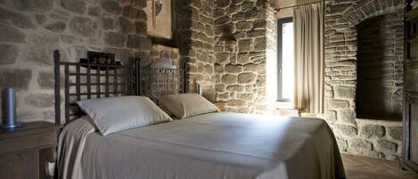 A stone-walled bedroom with a bed, a nightstand, and a window with curtains.