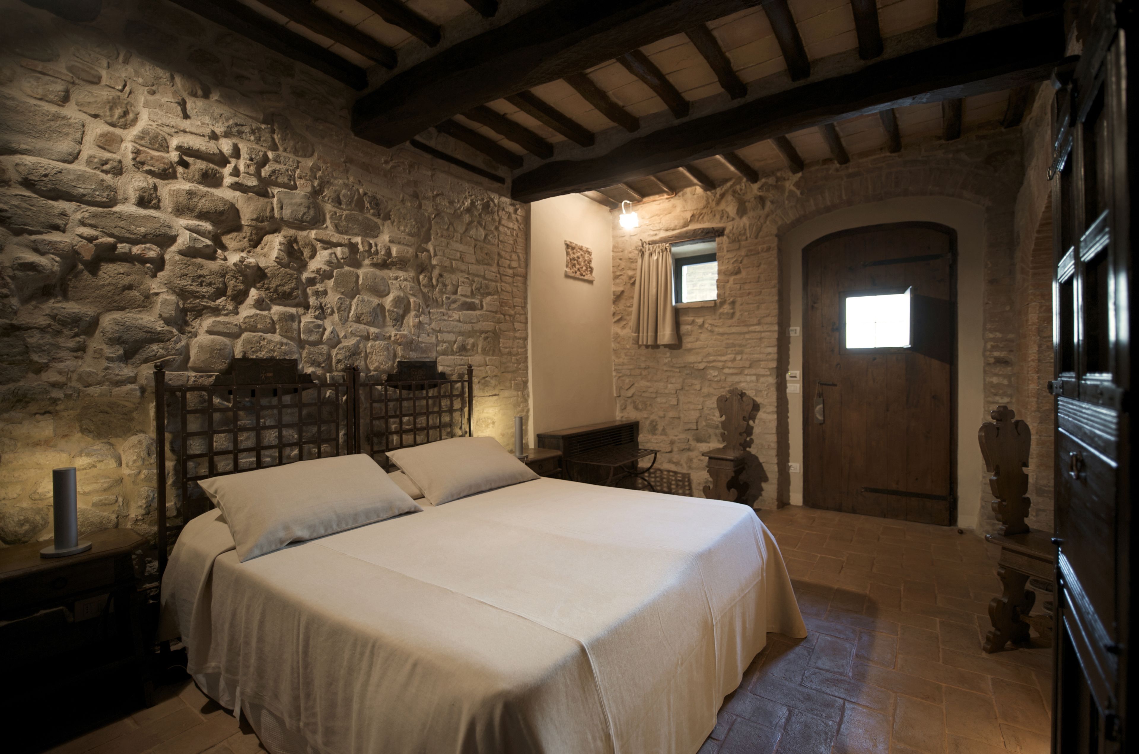 A bedroom with a stone wall, a wooden bed frame, a nightstand, and a small window with curtains.