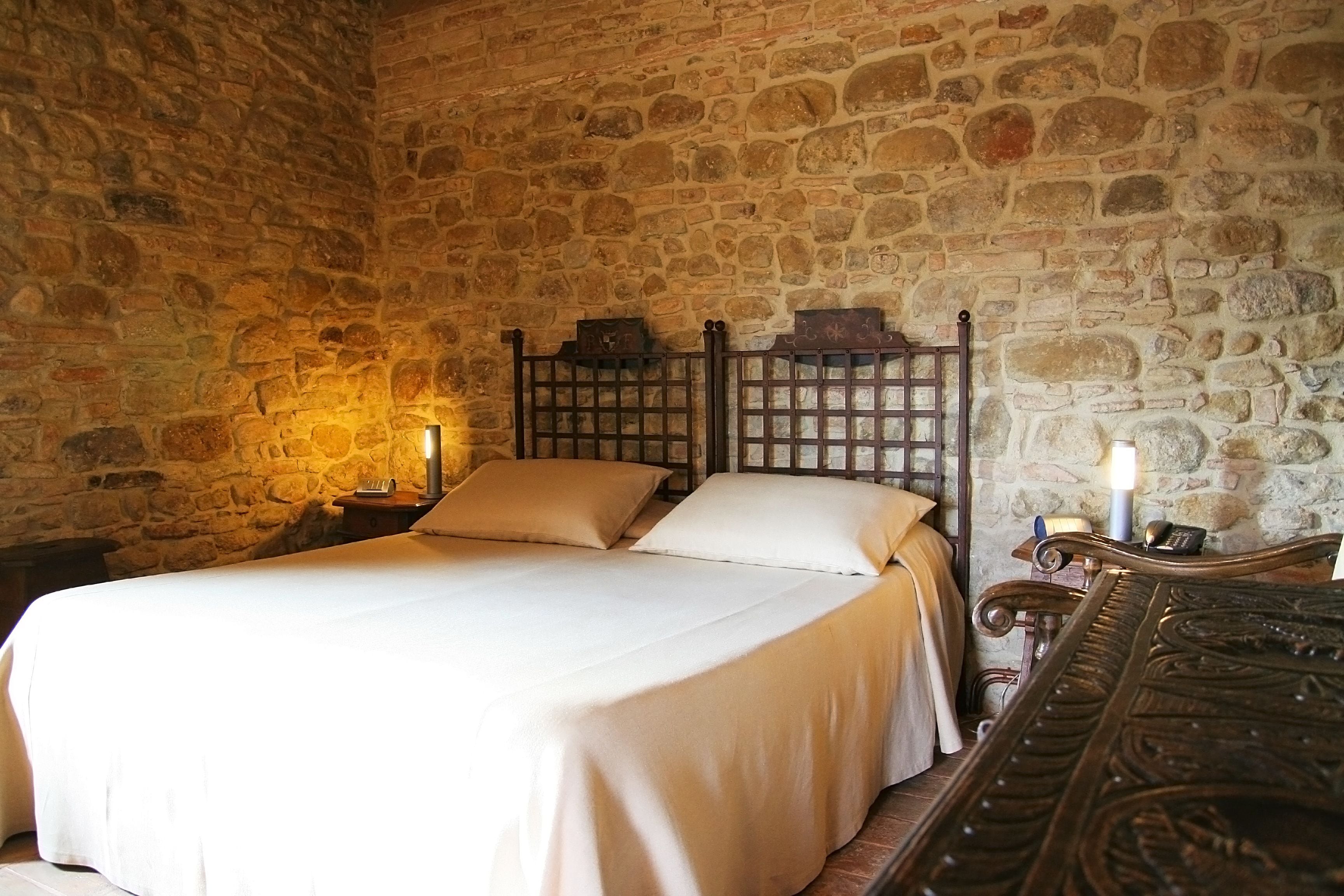 A room with a stone wall, a bed with a wooden headboard, and a nightstand with a lamp.