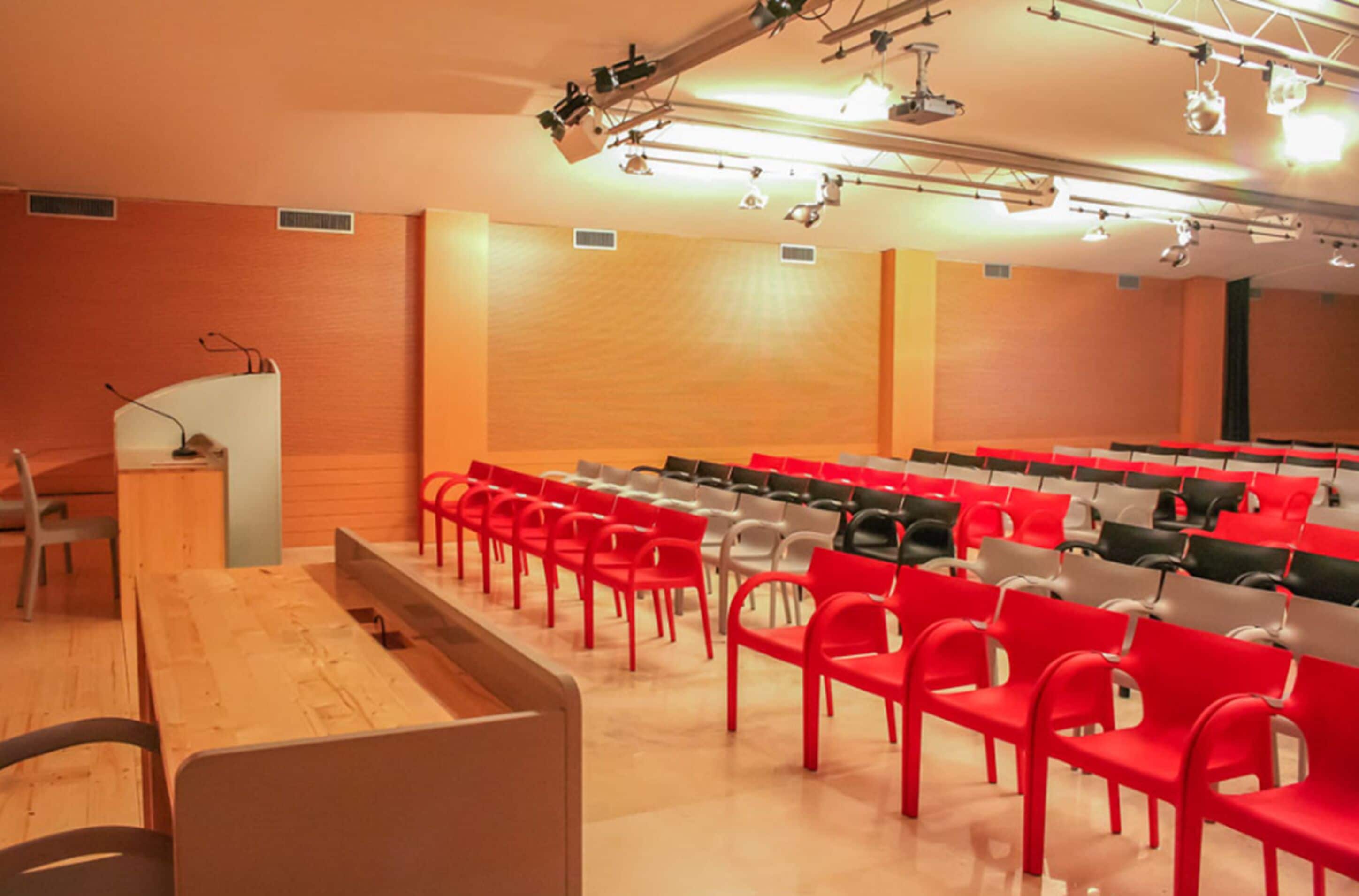 Une salle de conférence avec des rangées de chaises rouges et grises, une table en bois et un pupitre.