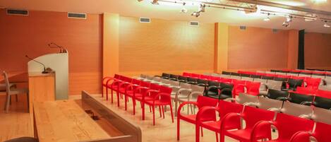 Une salle de conférence avec des rangées de chaises rouges et grises, une table en bois et un pupitre.