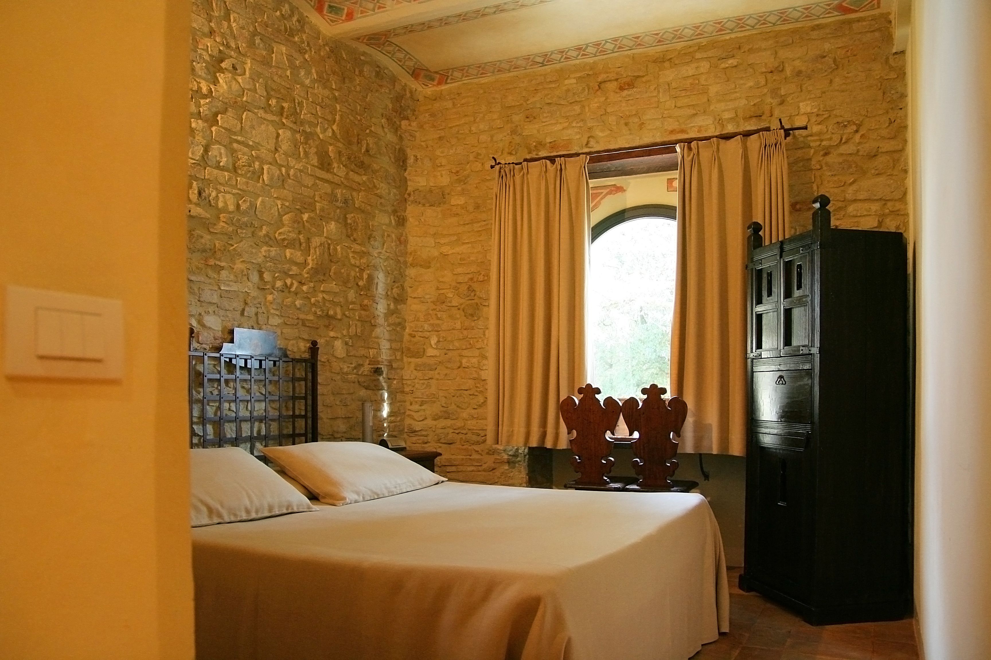 A bedroom with a stone wall, a bed with a beige bedspread, a wooden cabinet, and a window with curtains.