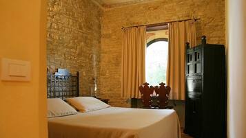 A bedroom with a stone wall, a bed with a beige bedspread, a wooden cabinet, and a window with curtains.