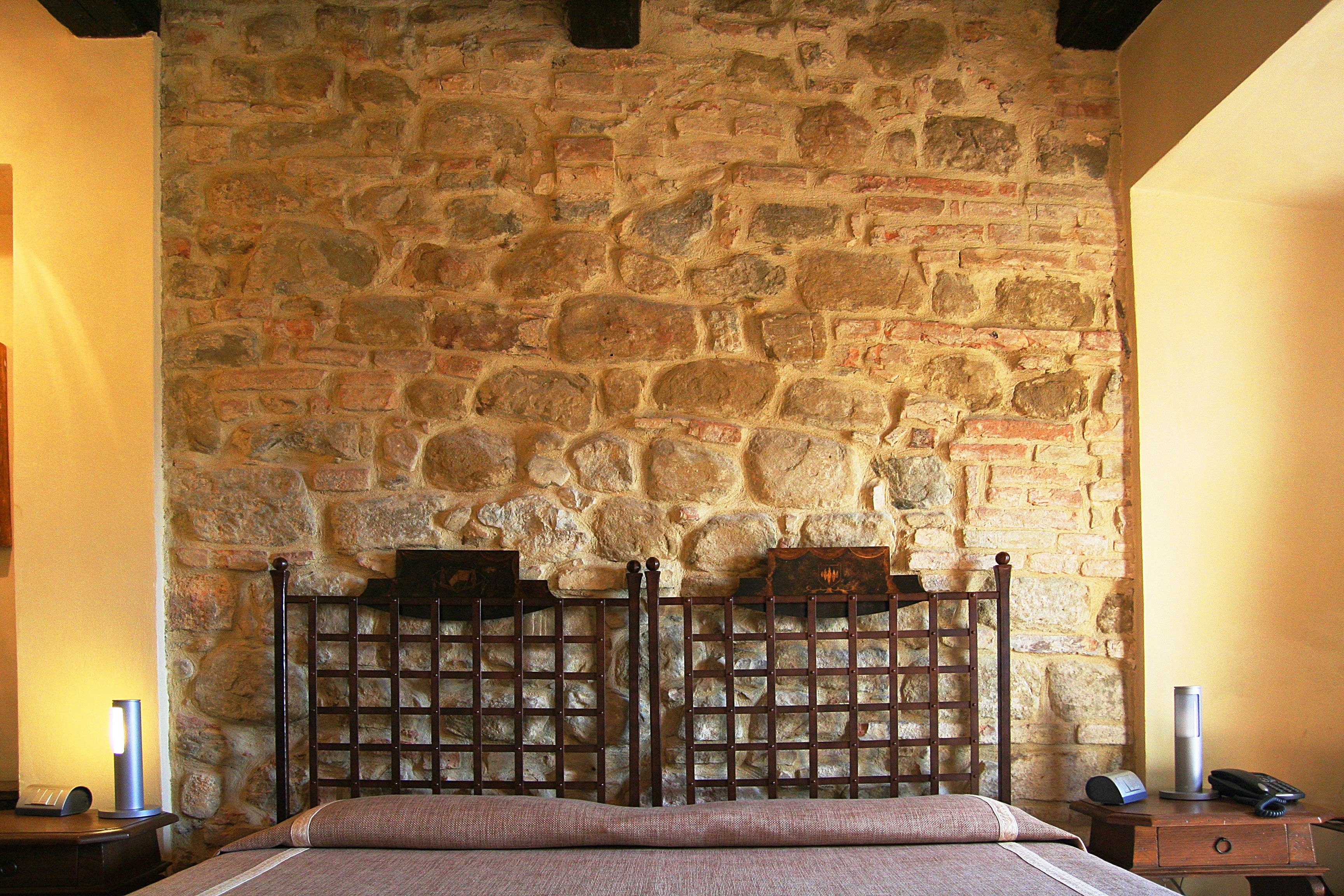 Una habitación con pared de piedra, estructura de cama de madera, mesita de noche con lámpara y mesita con teléfono.