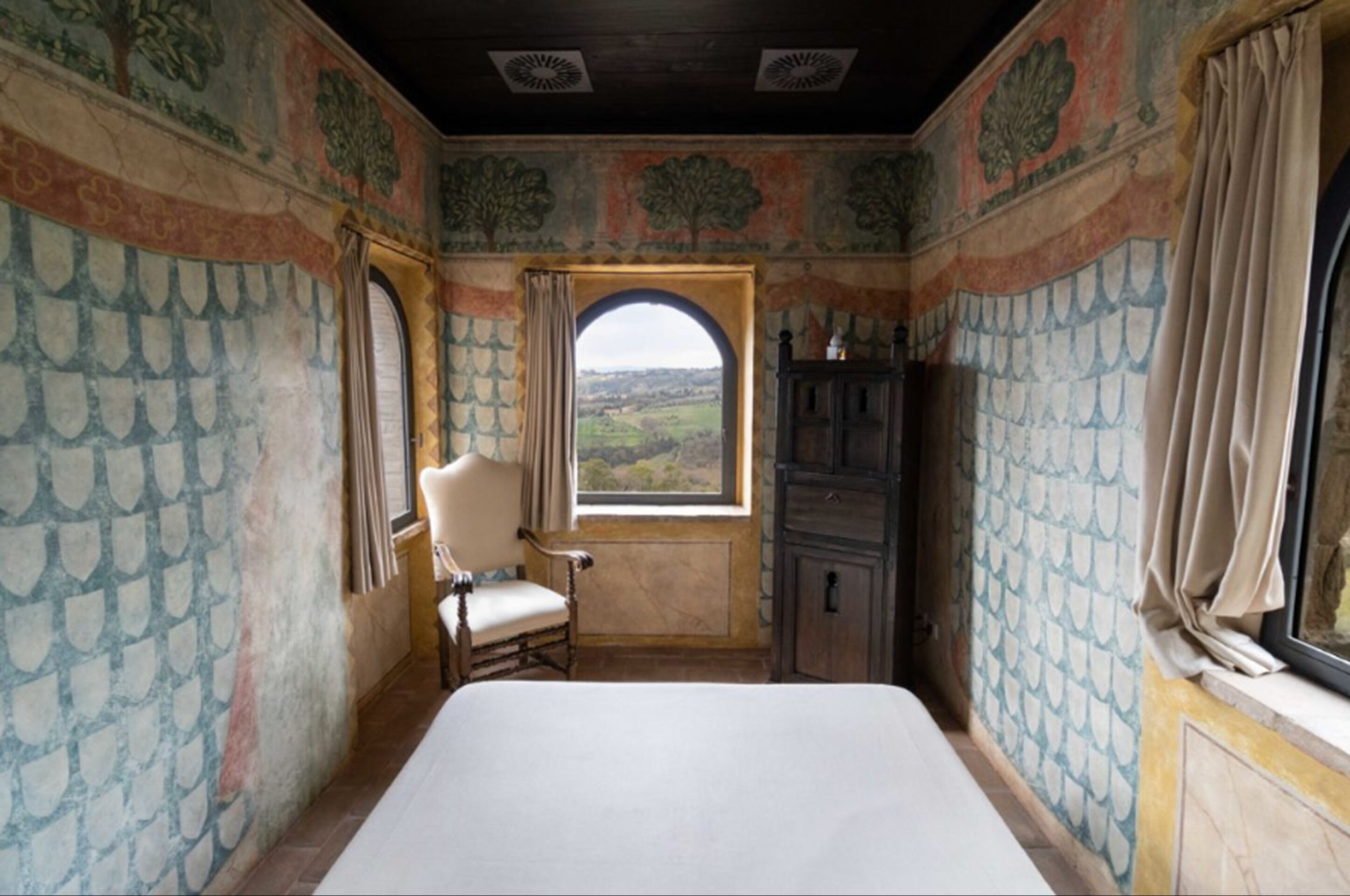 A room with a bed, a chair, a window with a view of the countryside, and a cabinet.