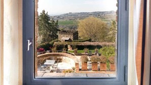 A view through an arched window of a courtyard with a stone building, a bridge, and greenery. - TH Perugia - Castello di Monterone (Perugia)