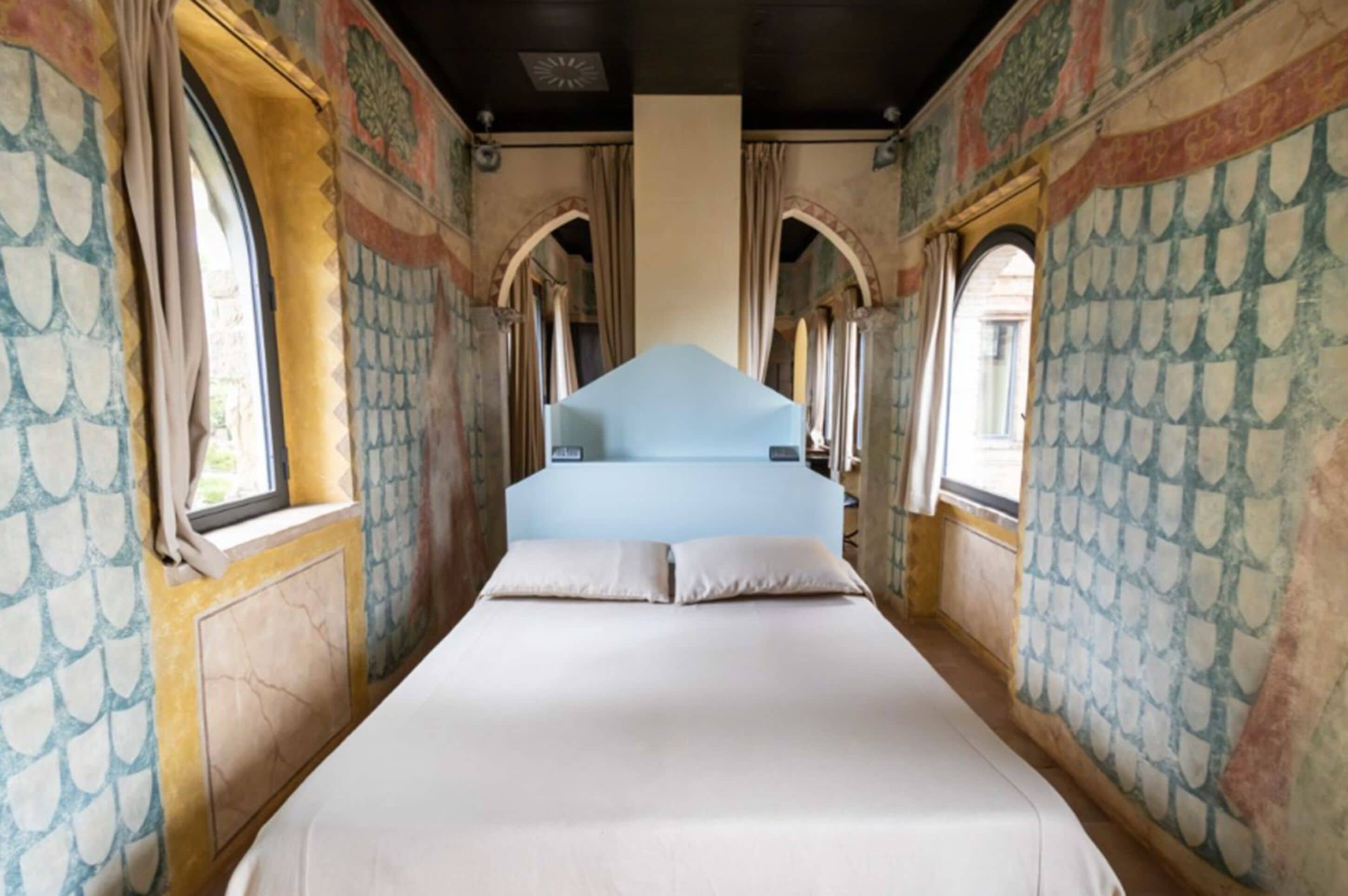 A narrow room with a bed centered in the middle, flanked by two windows with curtains. The walls have a patterned design.