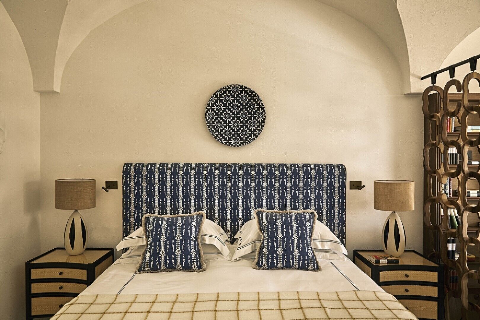 Un dormitorio con un cabecero estampado, dos mesitas de noche con lámparas, un adorno en la pared y una colcha con diseño.