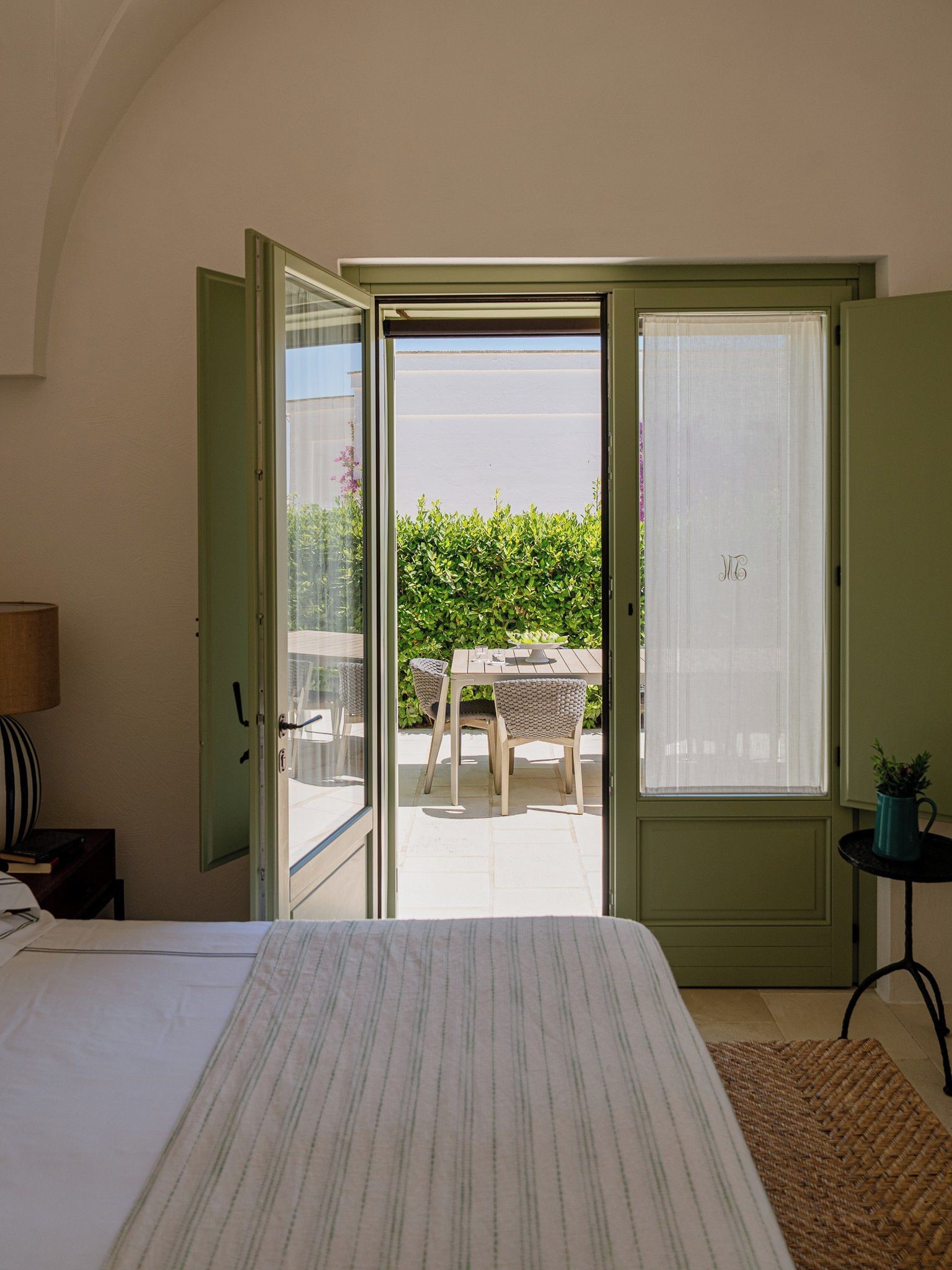 A bedroom with a bed, a nightstand, a lamp, and a view of an outdoor patio with a table and chairs.