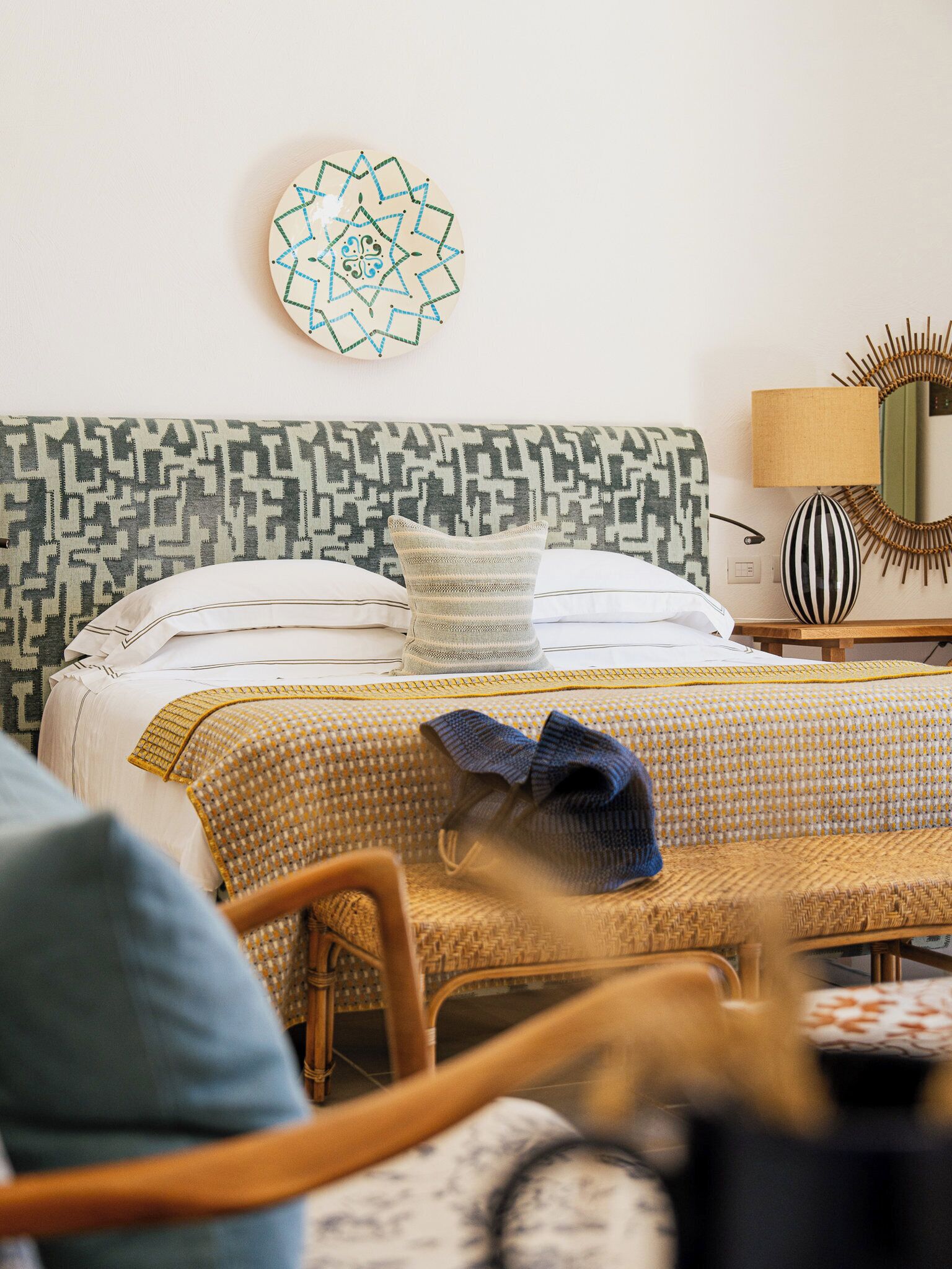 A neatly arranged bedroom with a patterned headboard, a decorative wall hanging, and a bedside table with a lamp.