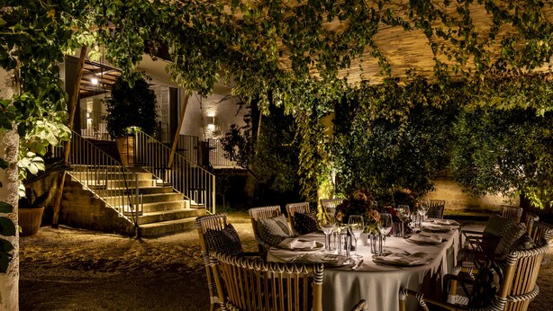 Un área de comedor al aire libre con sillas de mimbre y una mesa redonda preparada para una comida, rodeada de abundante vegetación y una escalera que conduce a un nivel superior.