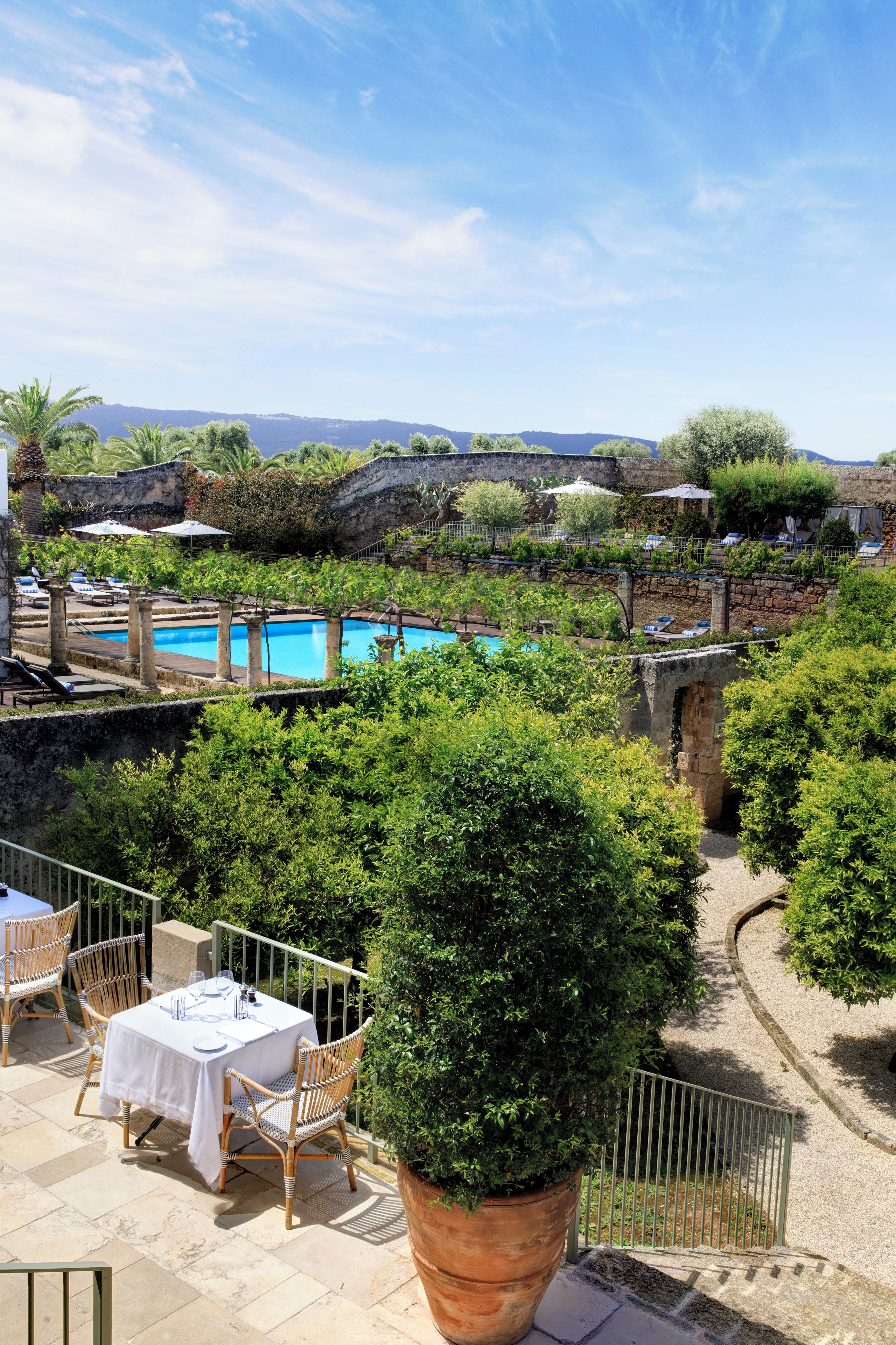 Zona de piscina con sillas de descanso y una mesa lista para comer, rodeada de follaje y muros de piedra.