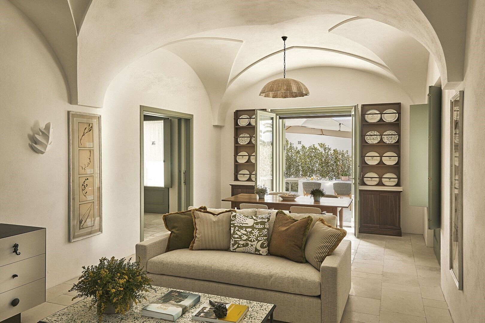 A spacious living area with a high arched ceiling, a sofa with patterned cushions, a coffee table with books and a plant, and a dining area with a view through a glass door.