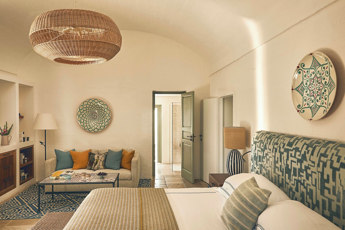 A well-lit, spacious room with a large bed, a sofa, a dining table, and a hanging light fixture.