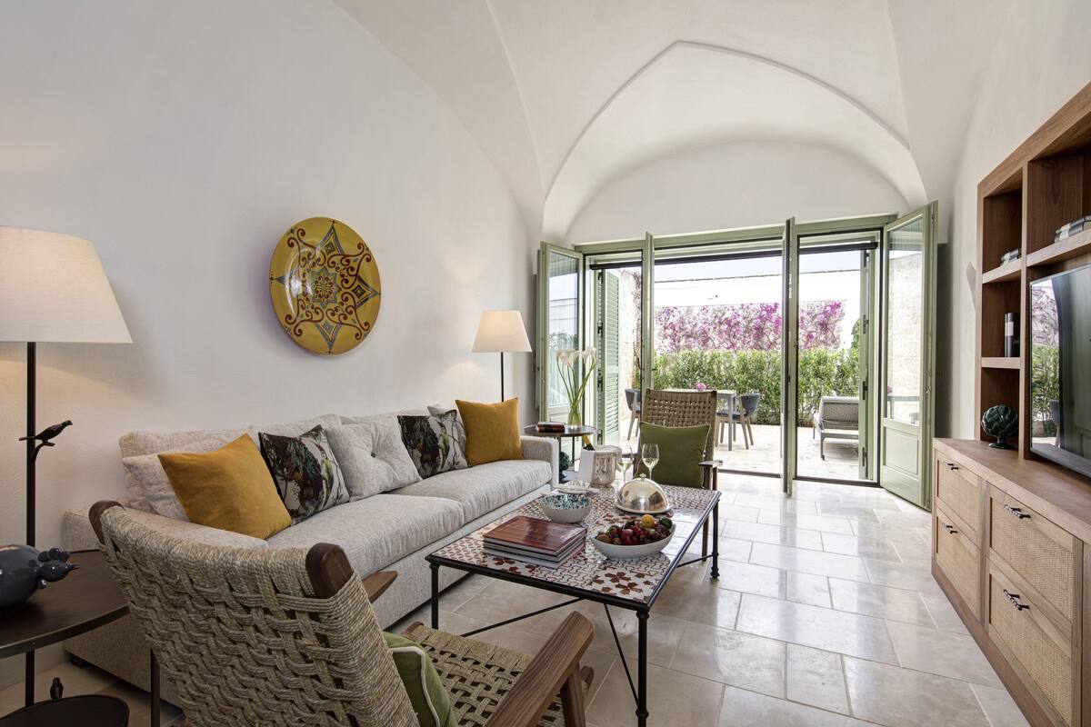 Ein modernes Wohnzimmer mit einer Couch, einem Couchtisch und Korbstühlen. Ein großes Fenster bietet einen Blick ins Freie.