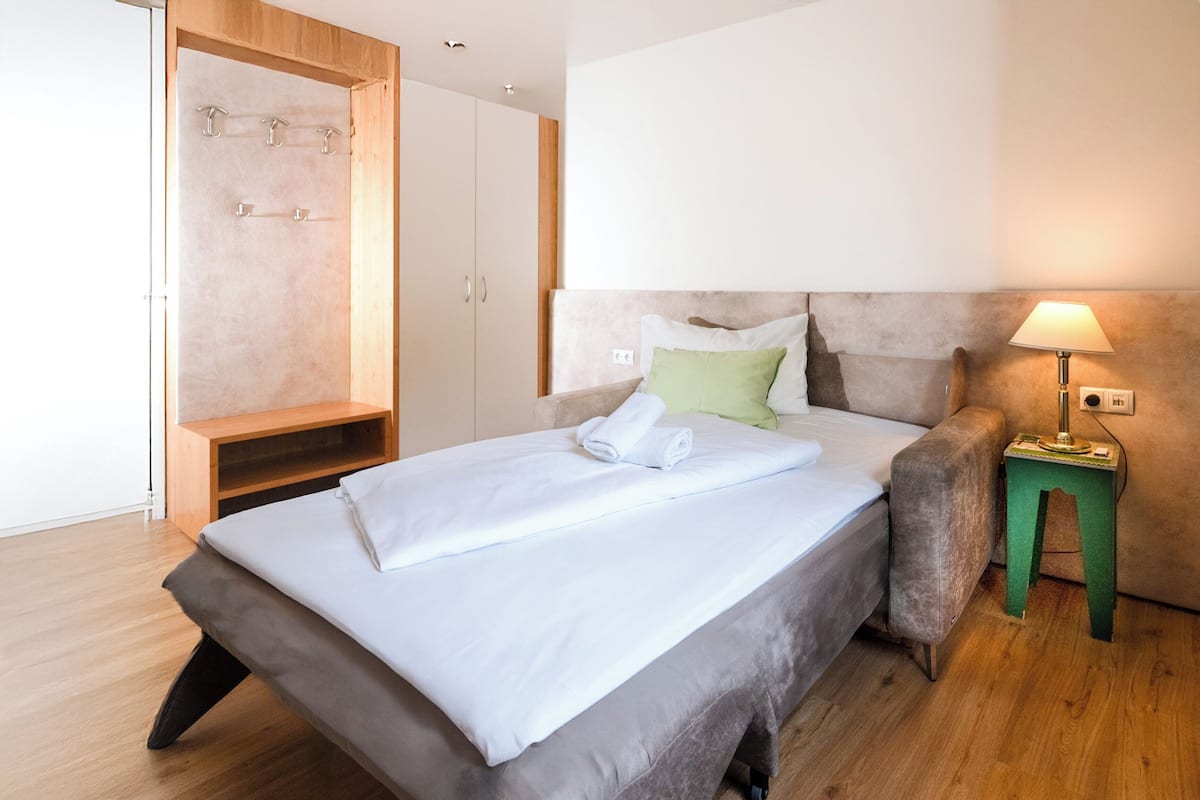 A modern bedroom with a bed, a nightstand, a lamp, a green stool, and a wardrobe.