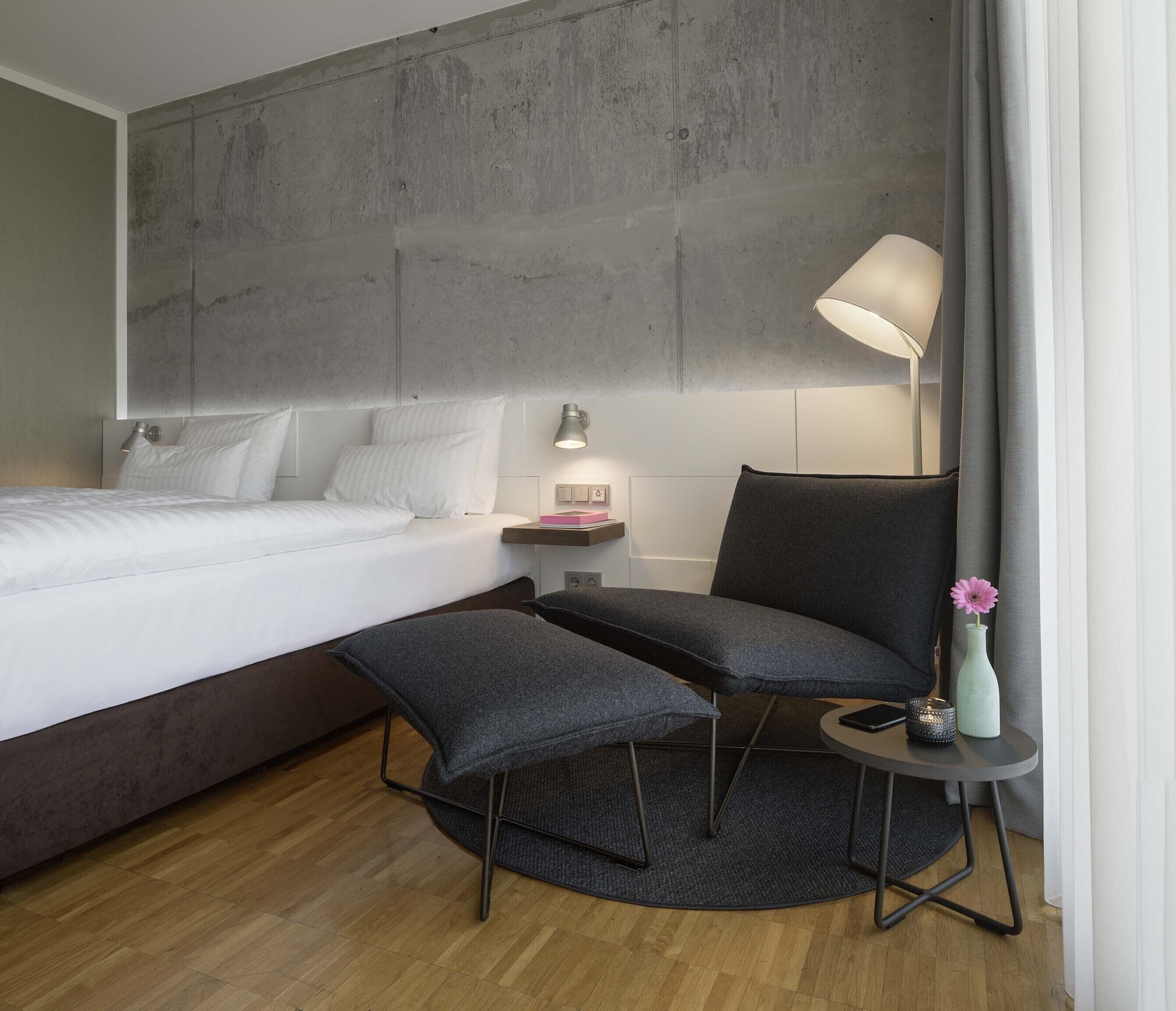 Ein modernes Hotelzimmer mit einem großen Bett, einem Schreibtisch mit Lampe, einem Sessel und einem kleinen Tisch mit Vase und Kerze.