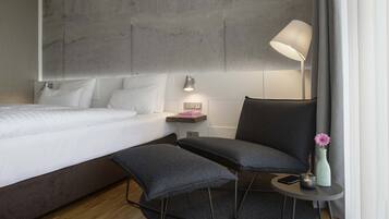Ein modernes Hotelzimmer mit einem großen Bett, einem Schreibtisch mit Lampe, einem Sessel und einem kleinen Tisch mit Vase und Kerze.