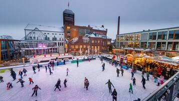 An outdoor ice skating rink surrounded by buildings with a prominent clock tower and a glass greenhouse structure.