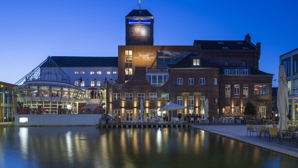 A waterfront hotel with a prominent tower, outdoor seating, and a glass facade restaurant. - Factory Hotel (Münster)