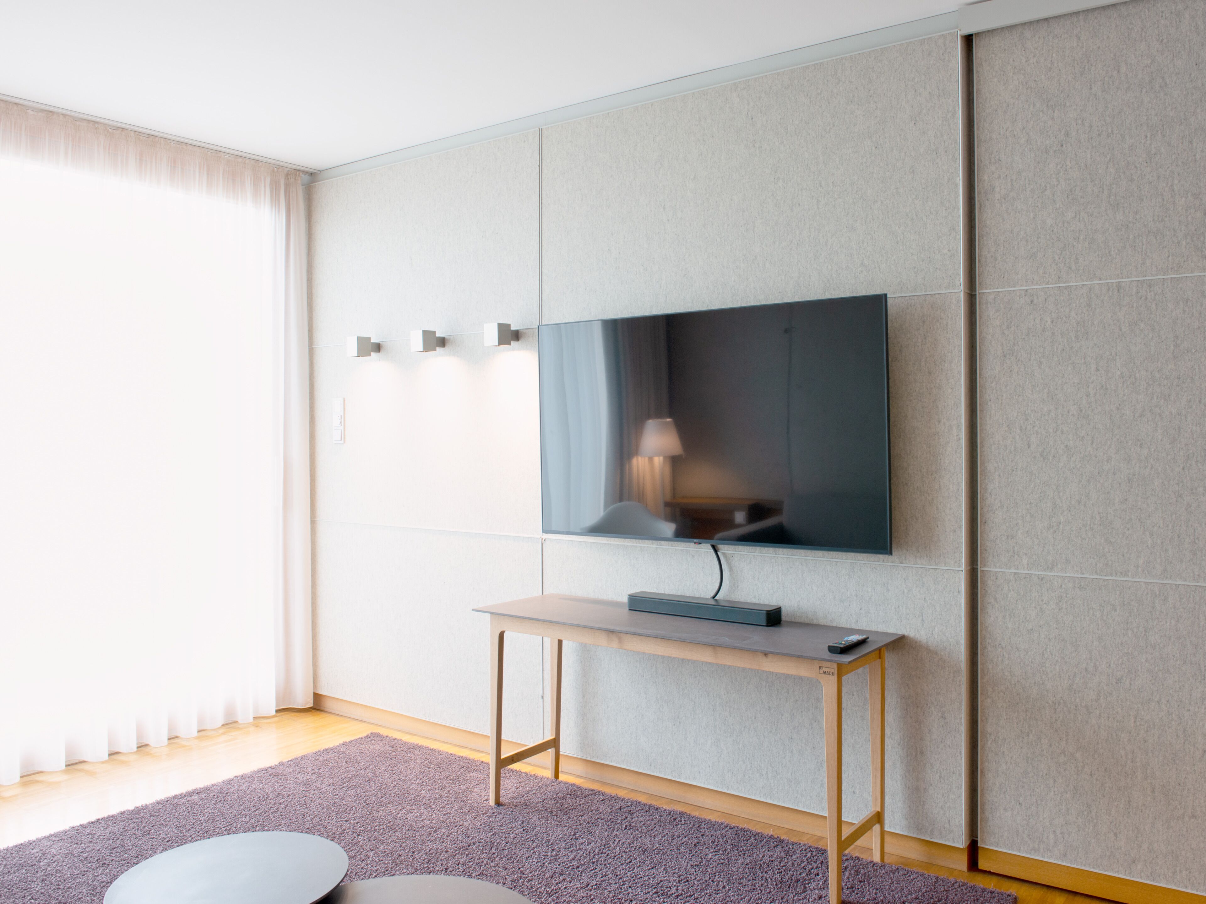 A modern hotel room with a large flat-screen TV mounted on a wall, a wooden side table, and a light-colored carpet.