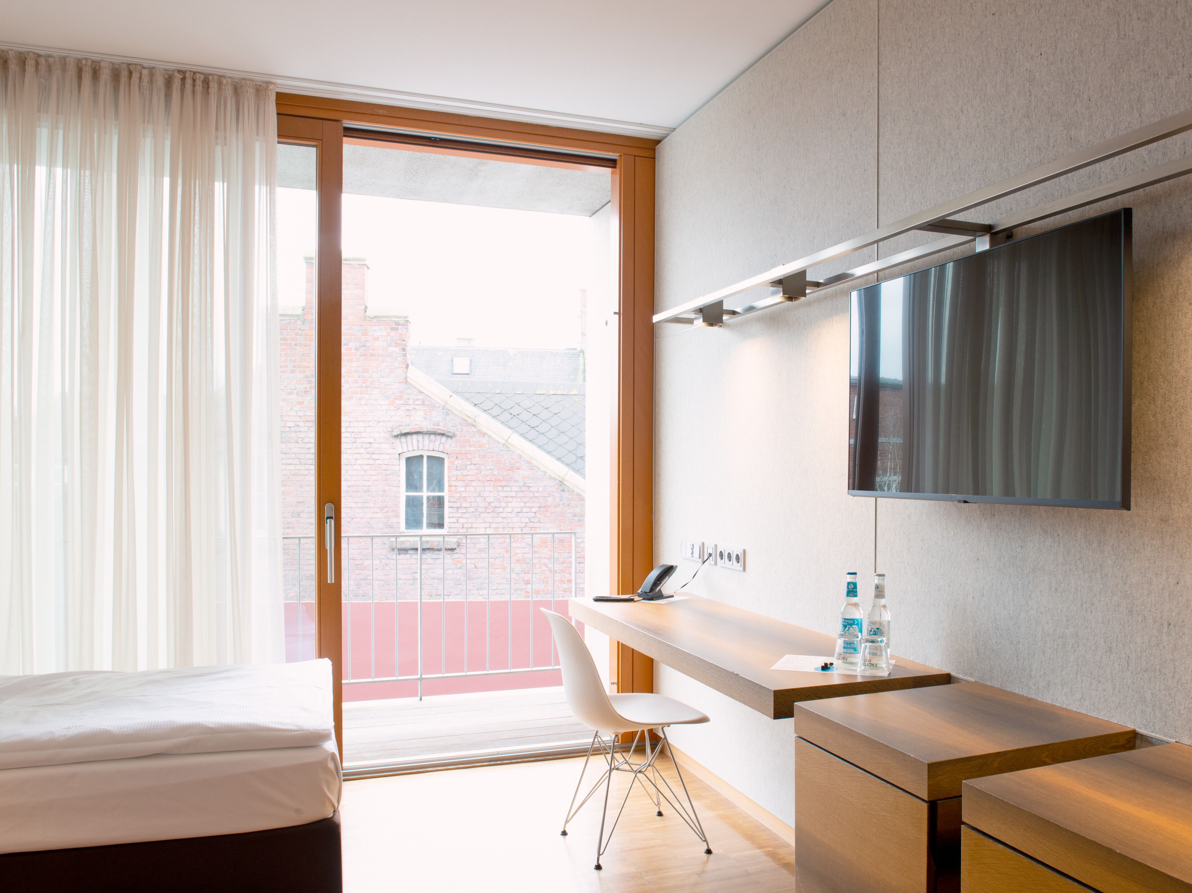 Een moderne hotelkamer met een bed, een bureau, een stoel, een flatscreen-tv en uitzicht op een bakstenen gebouw door een glazen schuifdeur.