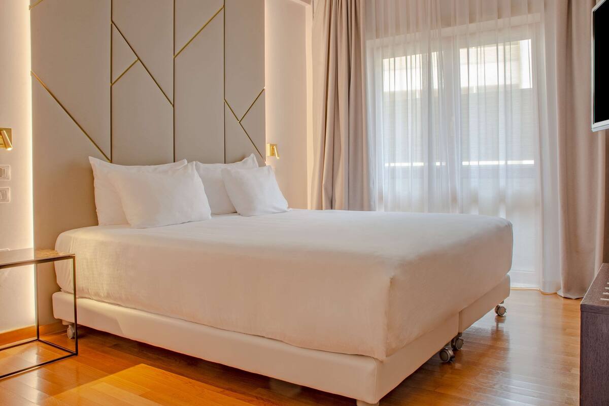 A modern bedroom with a large bed, a glass side table, a wall-mounted TV, and sheer curtains.