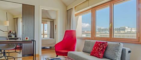 Una sala moderna con sofá gris, sillón rojo y ventanas amplias con vista a la ciudad.
