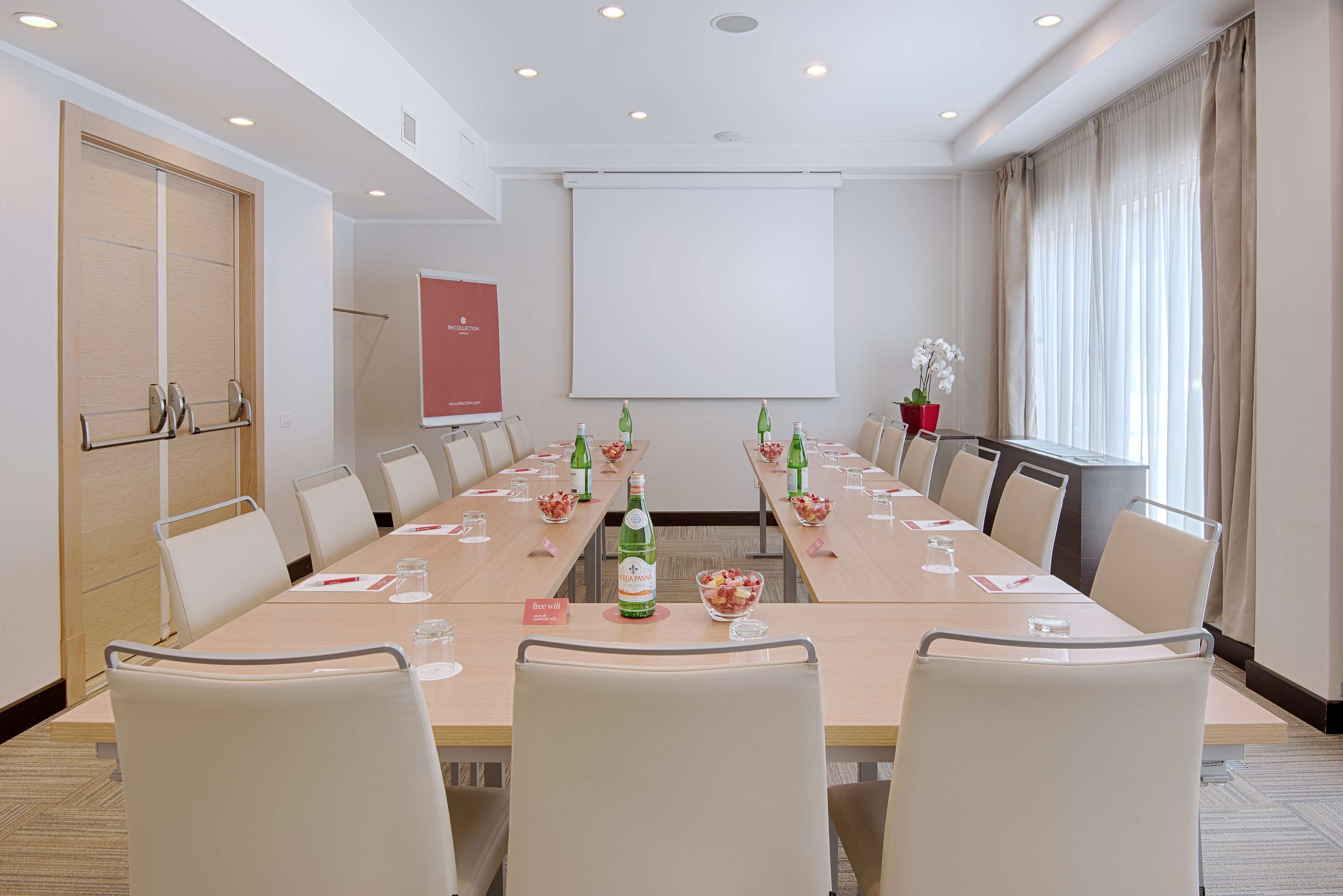 Un salón de conferencias con una larga mesa, sillas, una pantalla de proyector y un ventanal con cortinas.