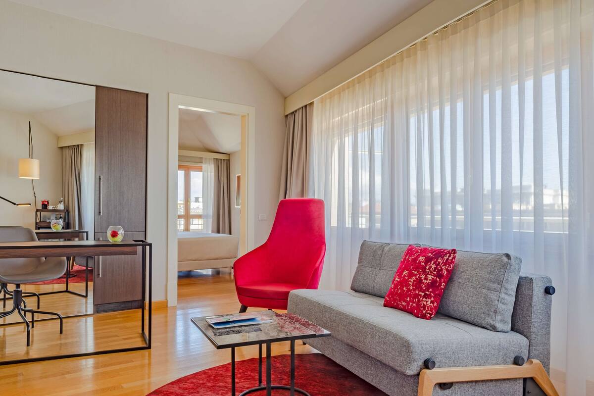 A modern living room with a grey sofa, red armchair, and a dining area with a wooden table and chairs.