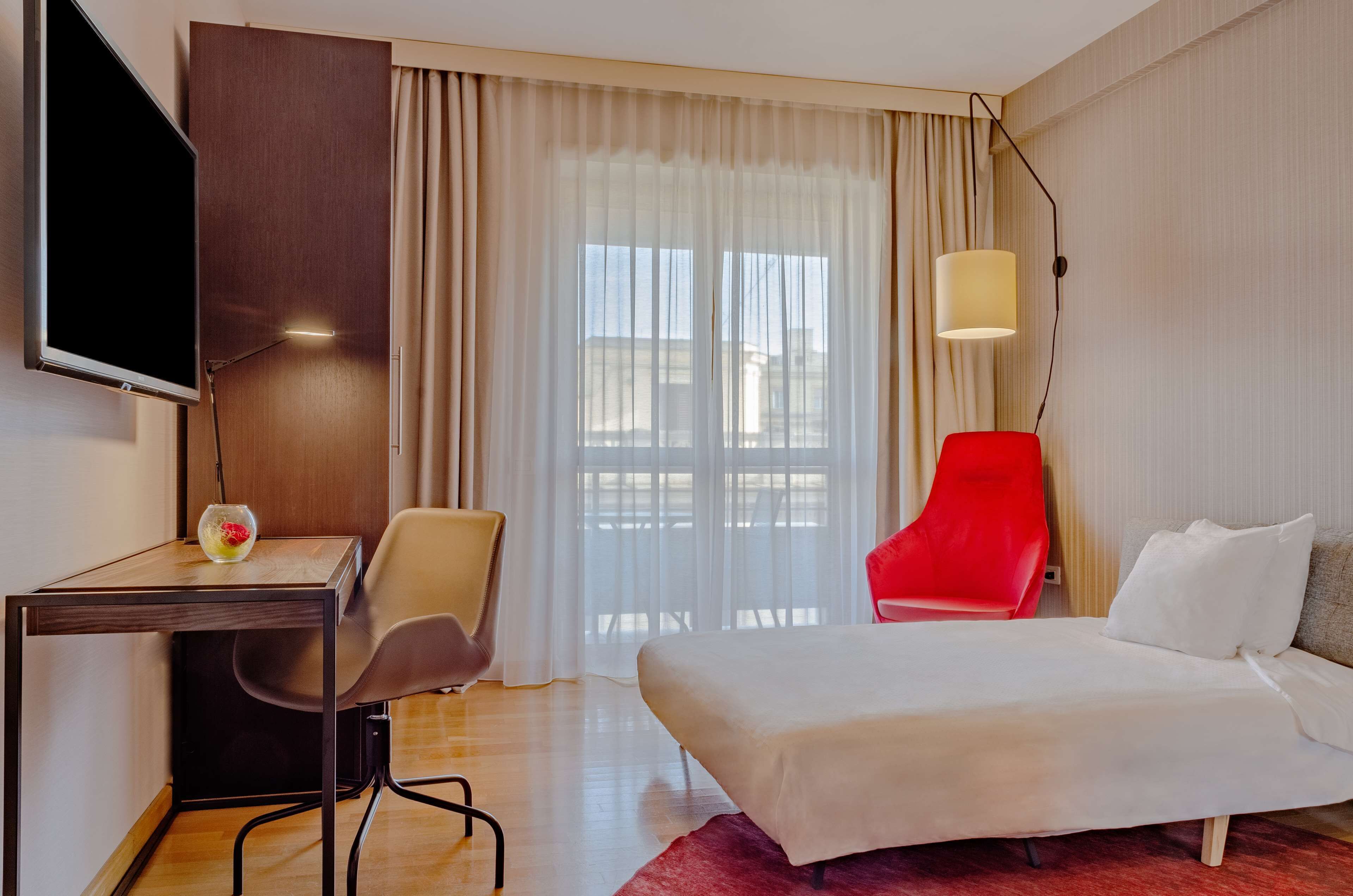 Una habitación de hotel con cama, escritorio, silla, televisión, una silla roja y una lámpara.