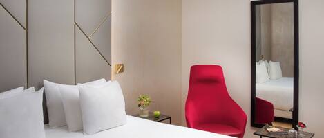 Habitación de hotel con cama, almohadas blancas, una silla roja, una mesita con un jarrón y una manzana, y un espejo grande que refleja la cama.