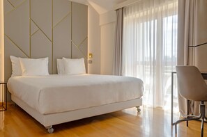 A modern hotel room with a large bed, a chair, and a view of the city through sheer curtains.
