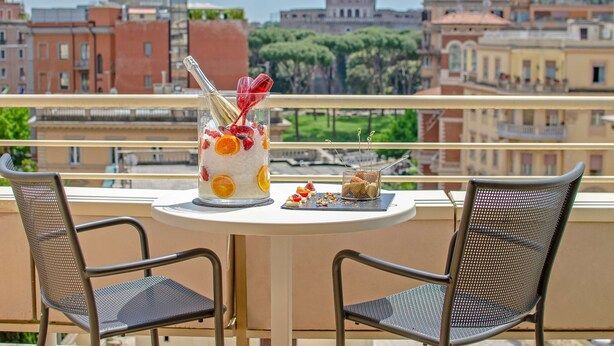 Een balkon met een ronde tafel en twee stoelen, een dessert met fruit en een glas, en uitzicht op stadsgebouwen en bomen.