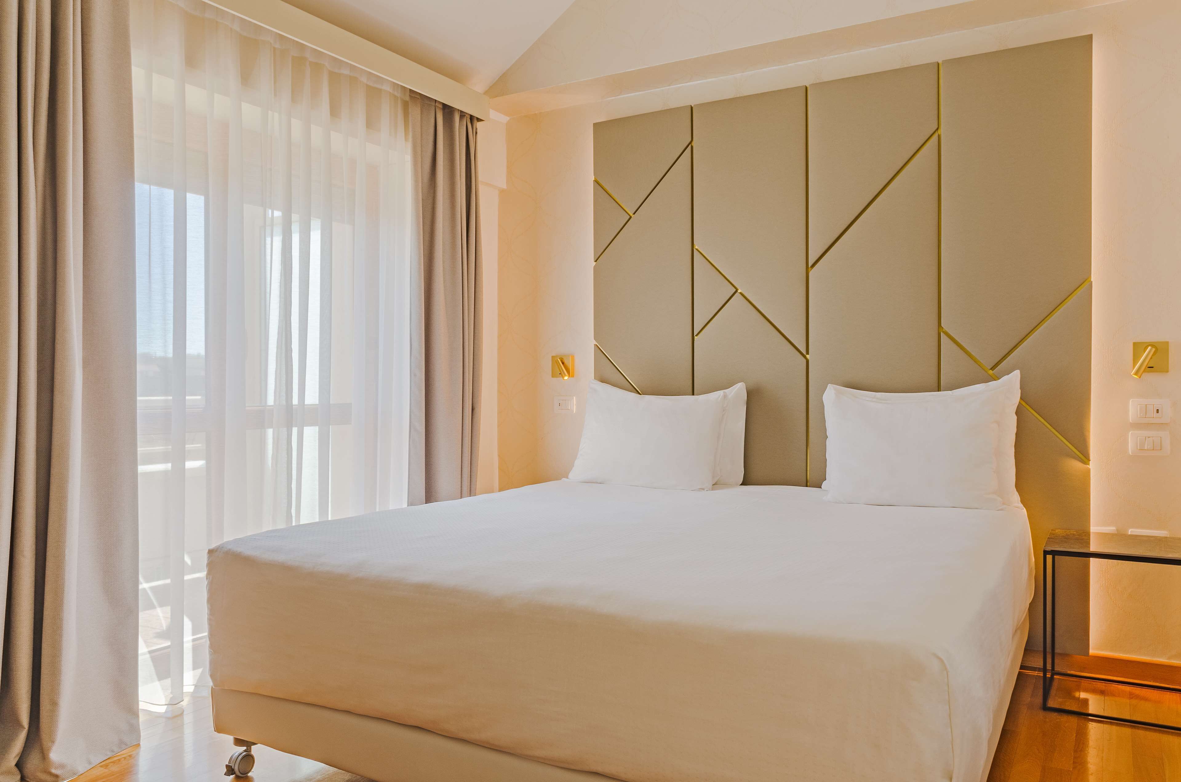 A hotel room with a large bed, a modern headboard, and a large window with curtains.