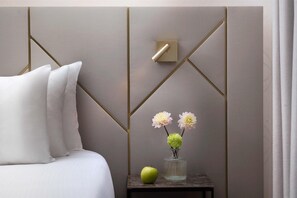 A neatly made bed with white linens, a small bedside table with a vase of flowers and an apple, and a geometric-patterned wall with a light switch.