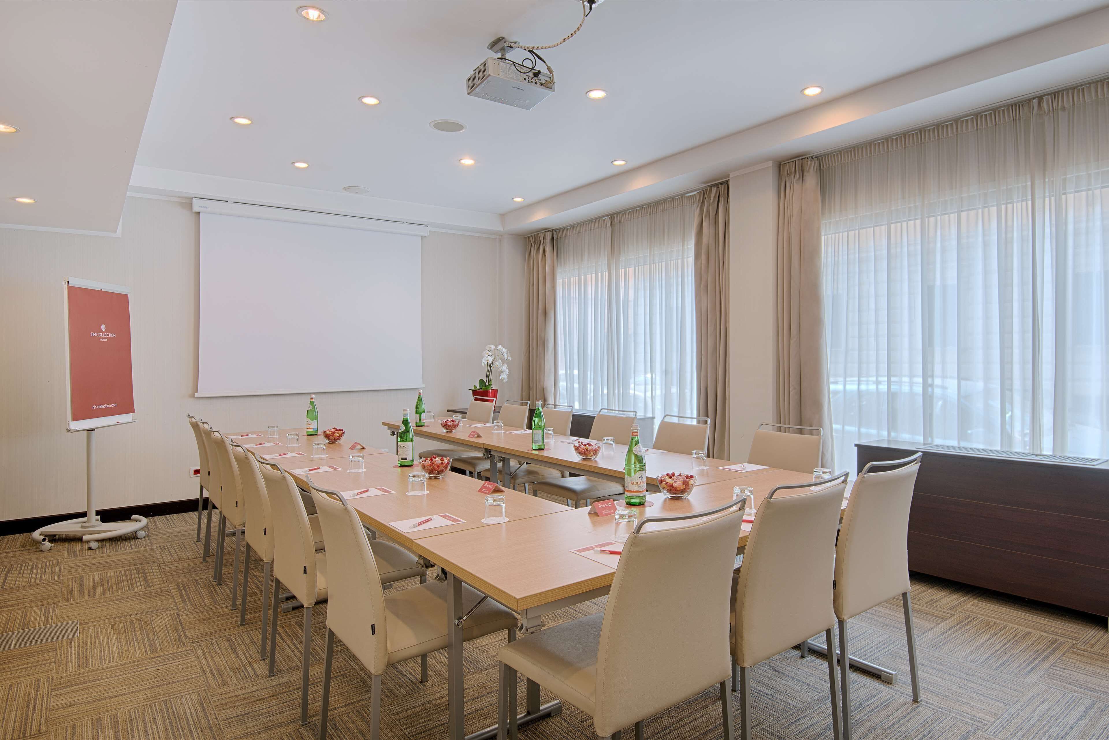A conference room with a long table, chairs, a projector, a whiteboard, and a large window with curtains.