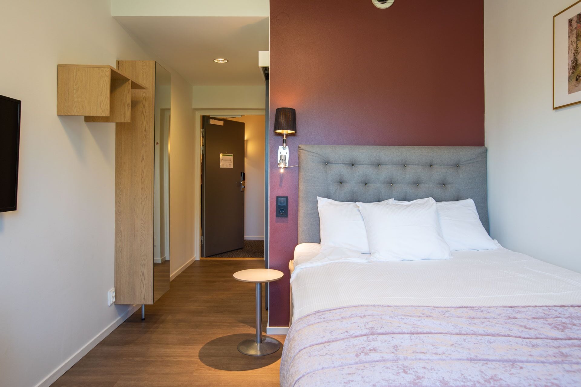 A hotel room with a bed, a bedside table, a lamp, and a wall-mounted TV.
