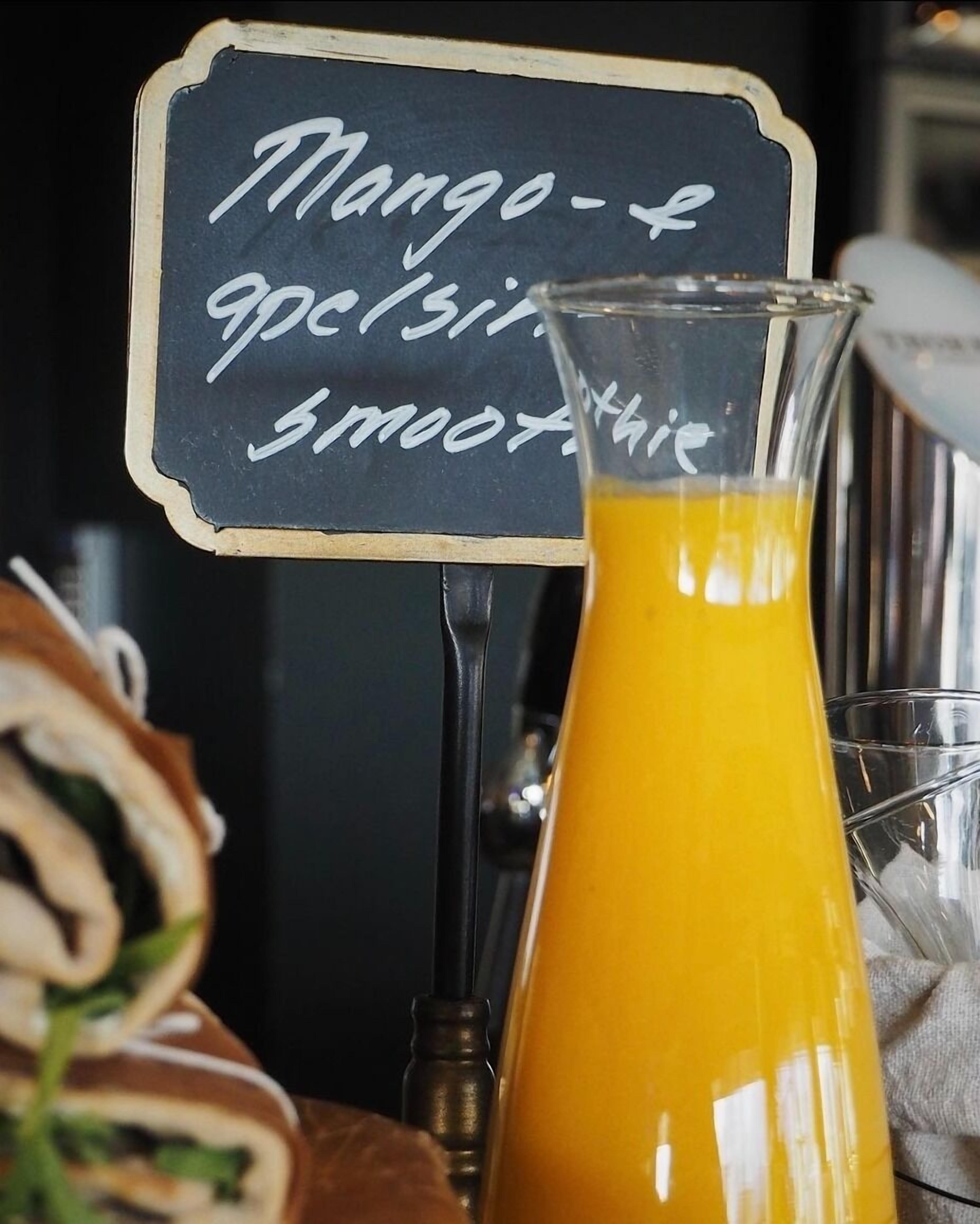 Une carafe en verre remplie de jus d’orange, avec un panneau indiquant qu’il y a aussi des smoothies à la mangue.