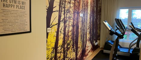 A gym with exercise equipment, a large forest mural, and a window with curtains.