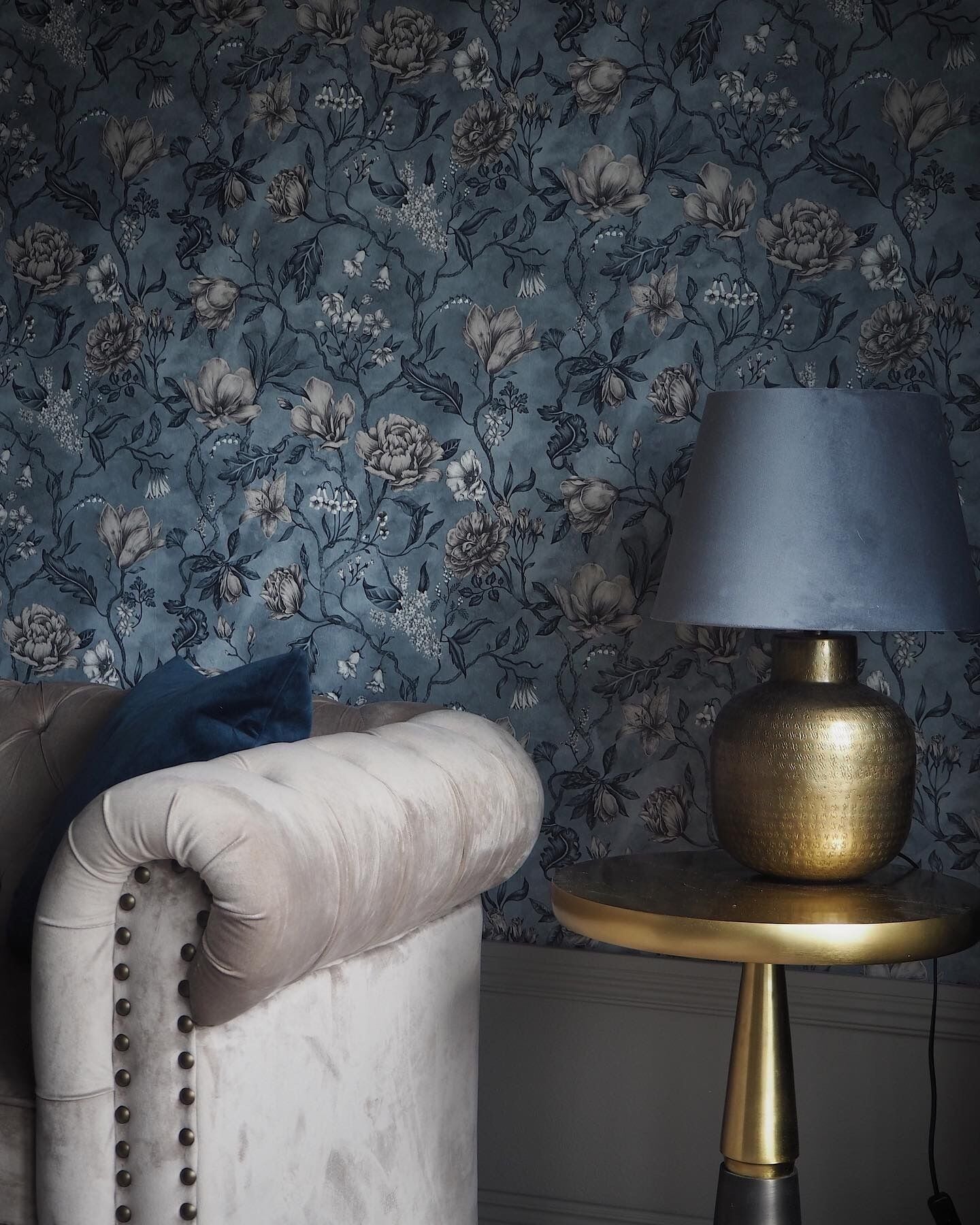 Et værelse med blomstret tapet, en hvid, quiltet lænestol, et rundt guld bord med en sort lampe og en blå pude.