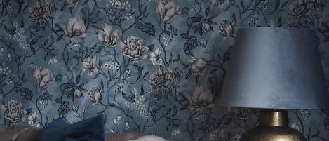 Una stanza con carta da parati a motivi floreali, una poltrona bianca con rivestimento trapuntato, un tavolino rotondo dorato con una lampada nera e un cuscino blu.