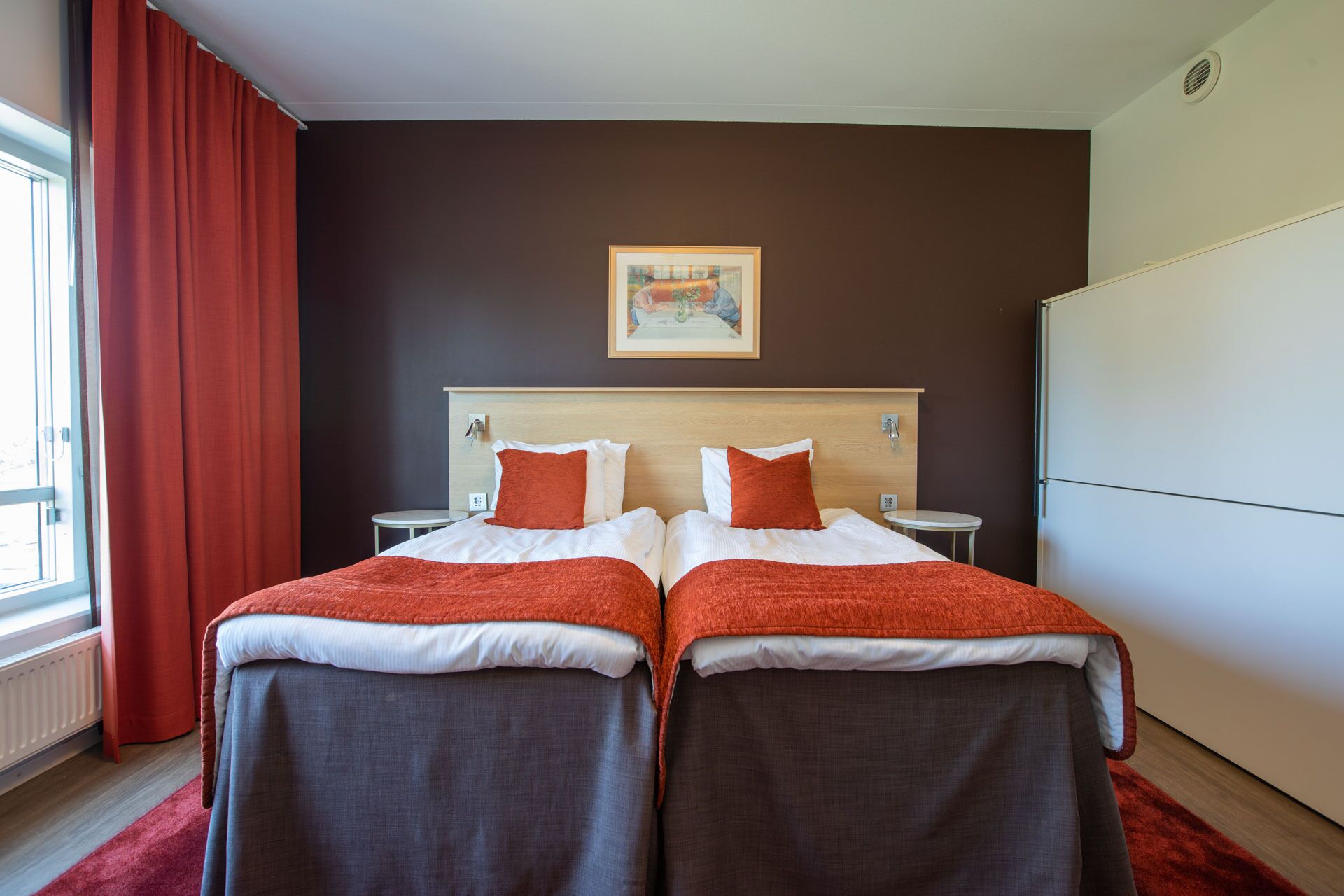 A double room with two beds, a painting on the wall, a white wardrobe, and a red curtain.