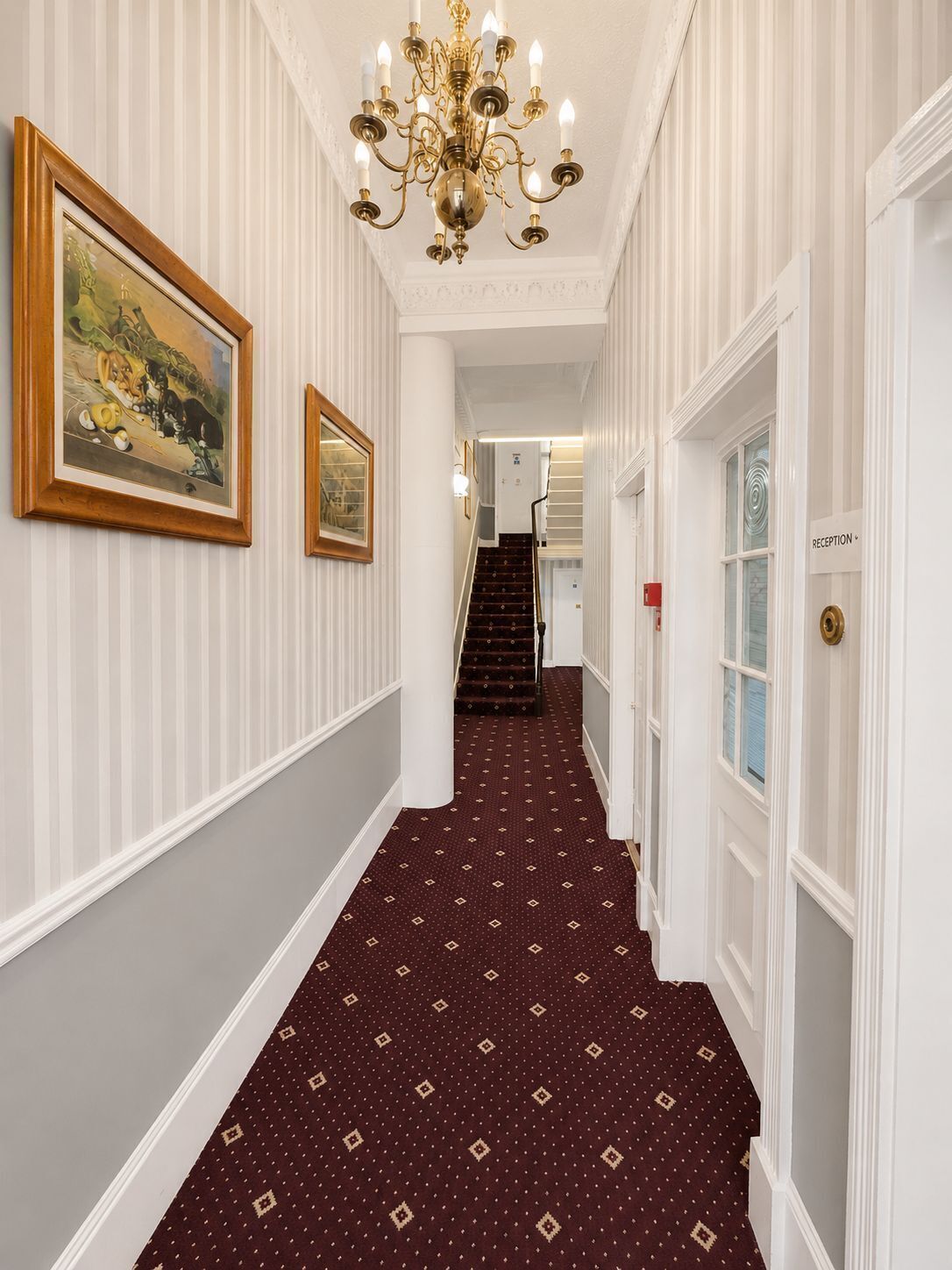 A hallway with a patterned carpet, a chandelier, and framed artwork on the walls.