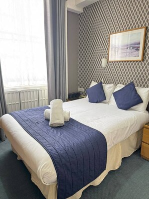 A hotel room with a large bed, a bedside table, a nightstand, a window with curtains, a patterned wall, and a framed picture on the wall. - Parkwood at Marble Arch (London)