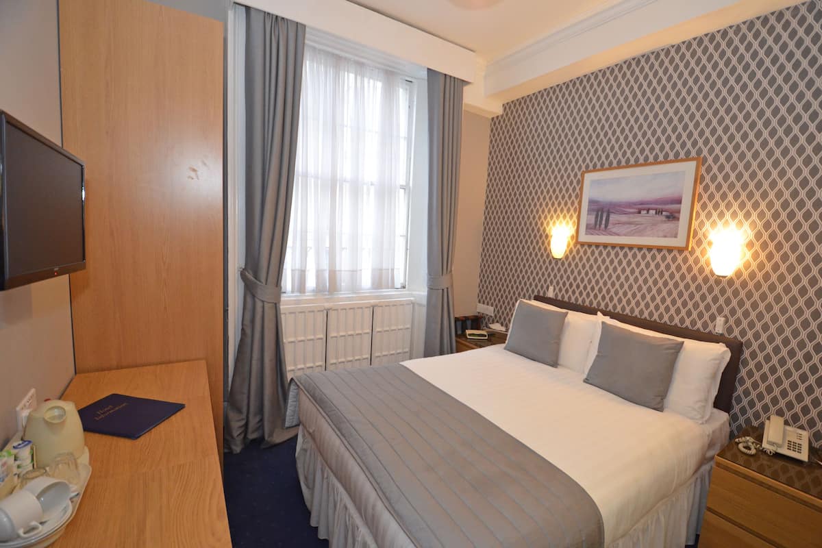 A hotel room with a large bed, a television, a bedside table with a lamp, a window with curtains, and a patterned wall.