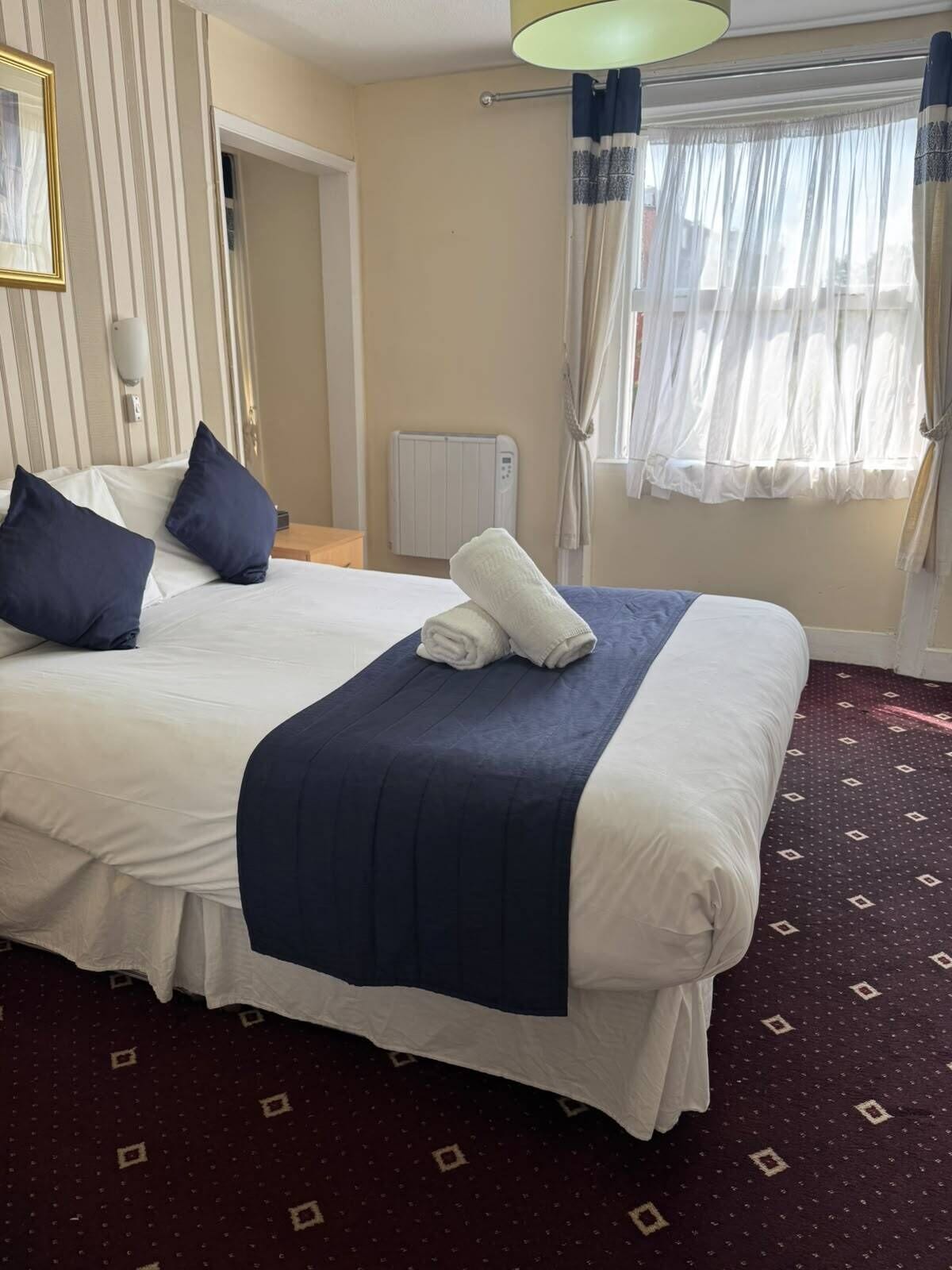 Una habitación de hotel con una cama grande, un radiador, una ventana con cortinas y una alfombra estampada.