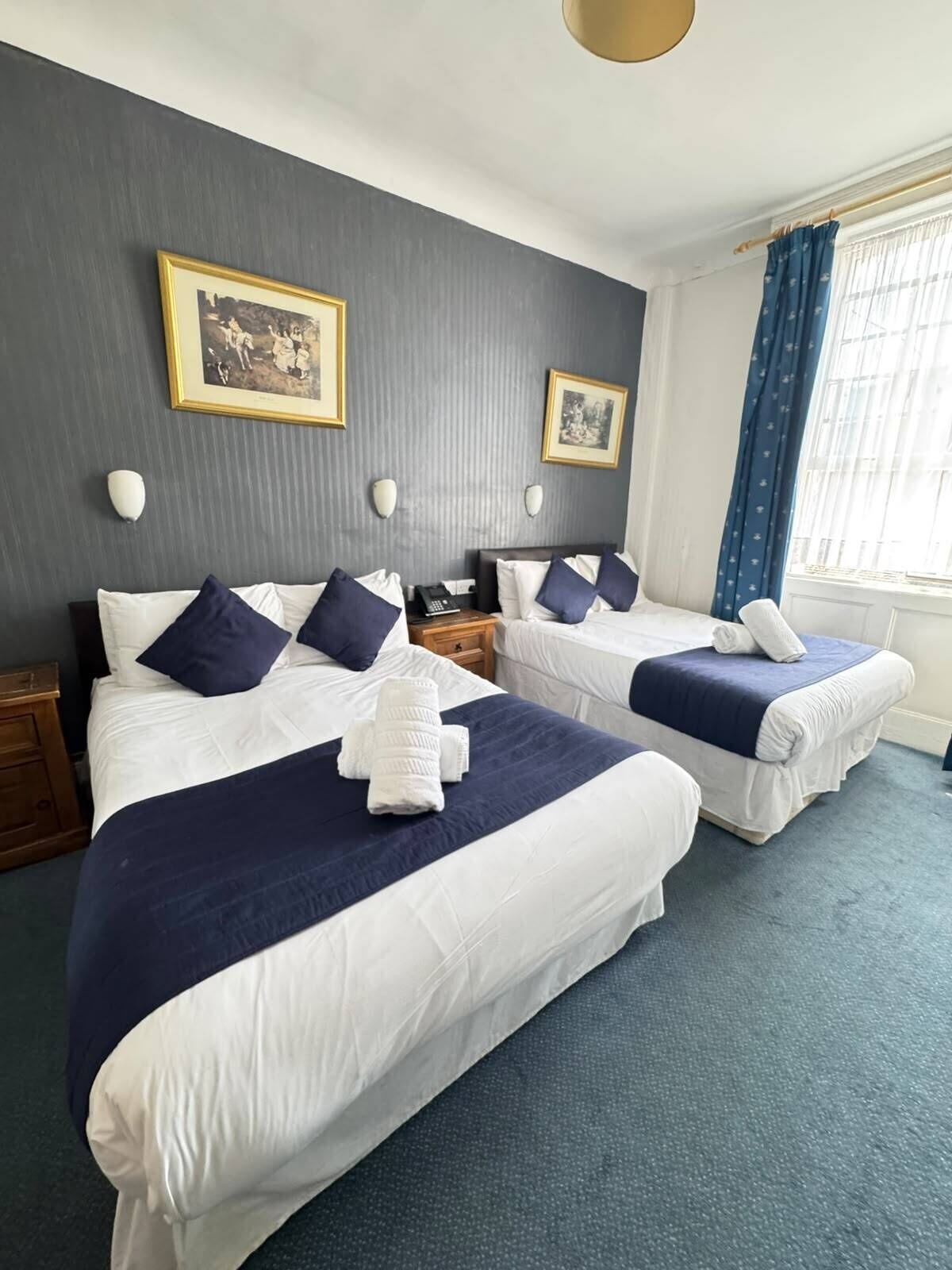 A hotel room with two beds, each with a blue and white duvet, a bedside table with a lamp, and a window with blue curtains.