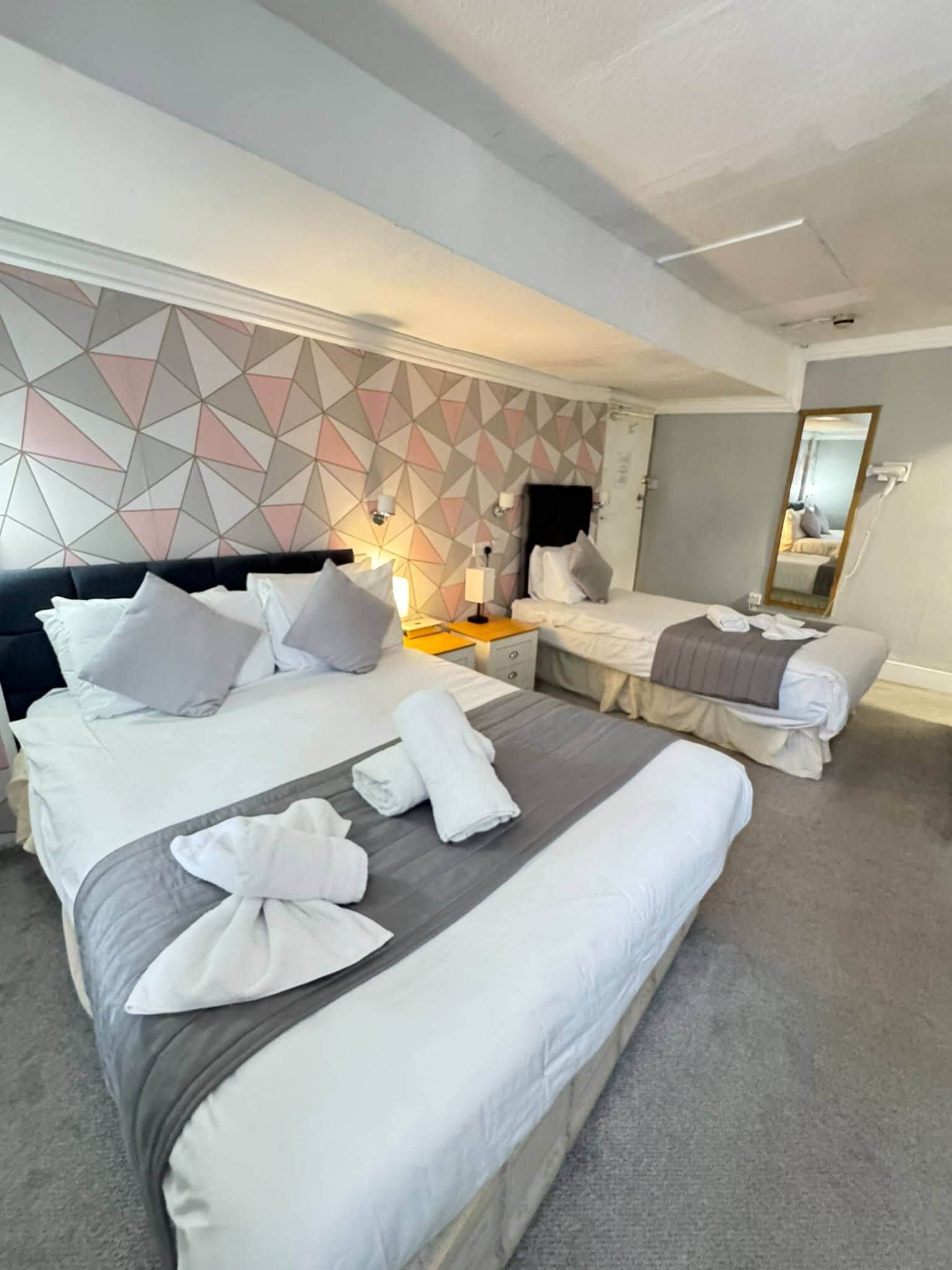 A hotel room with two beds, a geometric-patterned wall, a mirror, and a bedside table with a lamp.